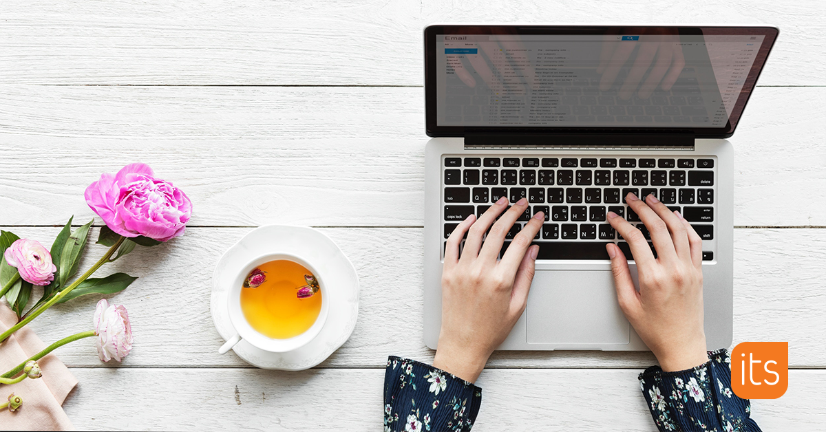 picture taken from above showing a desk with a laptop, a cup of tea and a pink flower. A female's hands are placed on the laptop's keyboard